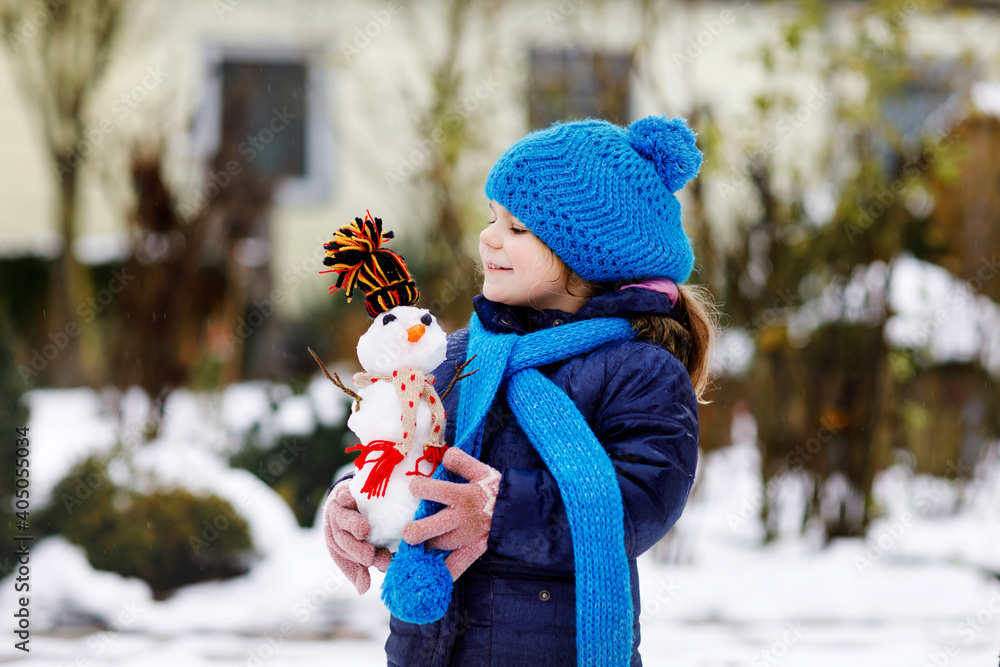 Cute little toddler girl making mini snowman and eating carrot nose. Adorable healthy happy child playing and having fun with snow, outdoors on cold day. Active leisure with children in winter