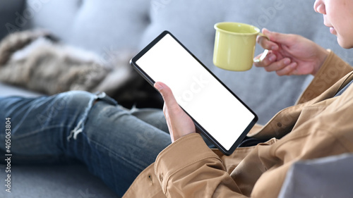 Close up view of young man using digital tablet and holding a cup of coffee while resting on sofa. © Prathankarnpap