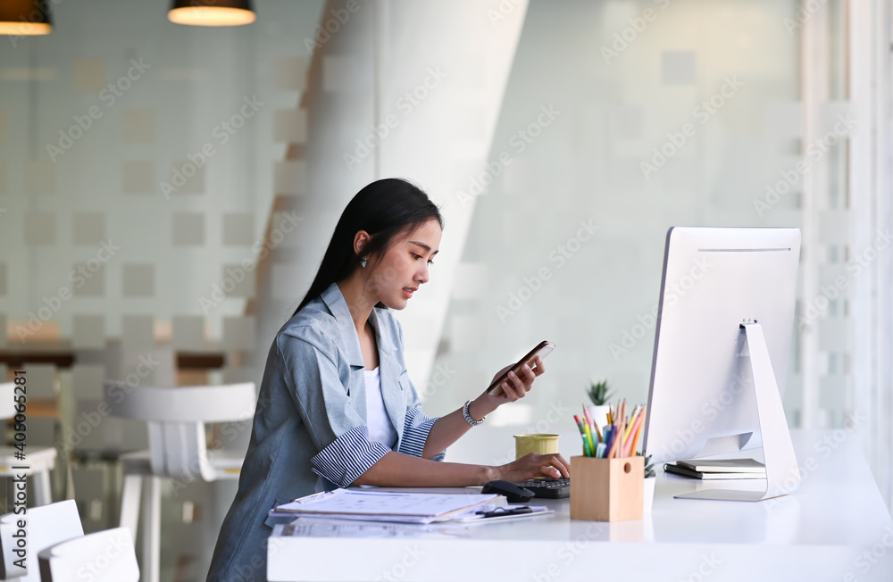 Side view of businesswoman working with computer and using smart phone while sitting in modern office. Side view of businesswoman working with computer and using smart phone while sitting in modern office.