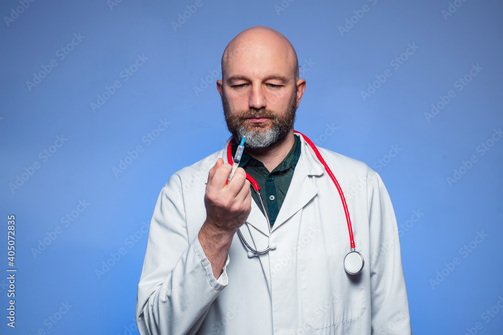Studio shot of a bald doctor with beard in his 40s, holding syringe in his hand, blue background. Vaccination and health care concept. Studio shot of a bald doctor with beard in his 40s, holding syringe in his hand, blue background. Vaccination and health care concept.