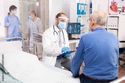 Medical staff questioning senior man in clinic consultation room wearing visor in the course of covid19. Physician practitioner in the course of private modern clinic appointment during coronavirus
