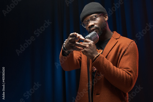 portrait of a dark-skinned handsome guy in brown jacket and black hat stands with a microphone and emotionally sings in a studio on dark background