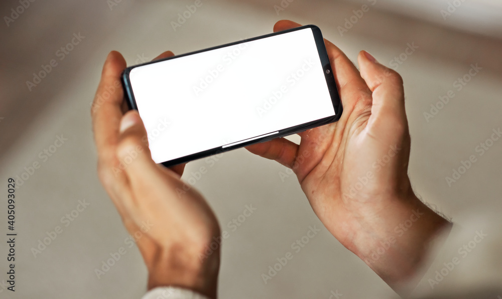 Closeup mockup image with blurred background of a man's hands holding modern mobile phone with blank white screen in a cozy room. Closeup mockup image with blurred background of a man's hands holding modern mobile phone with blank white screen in a cozy room.