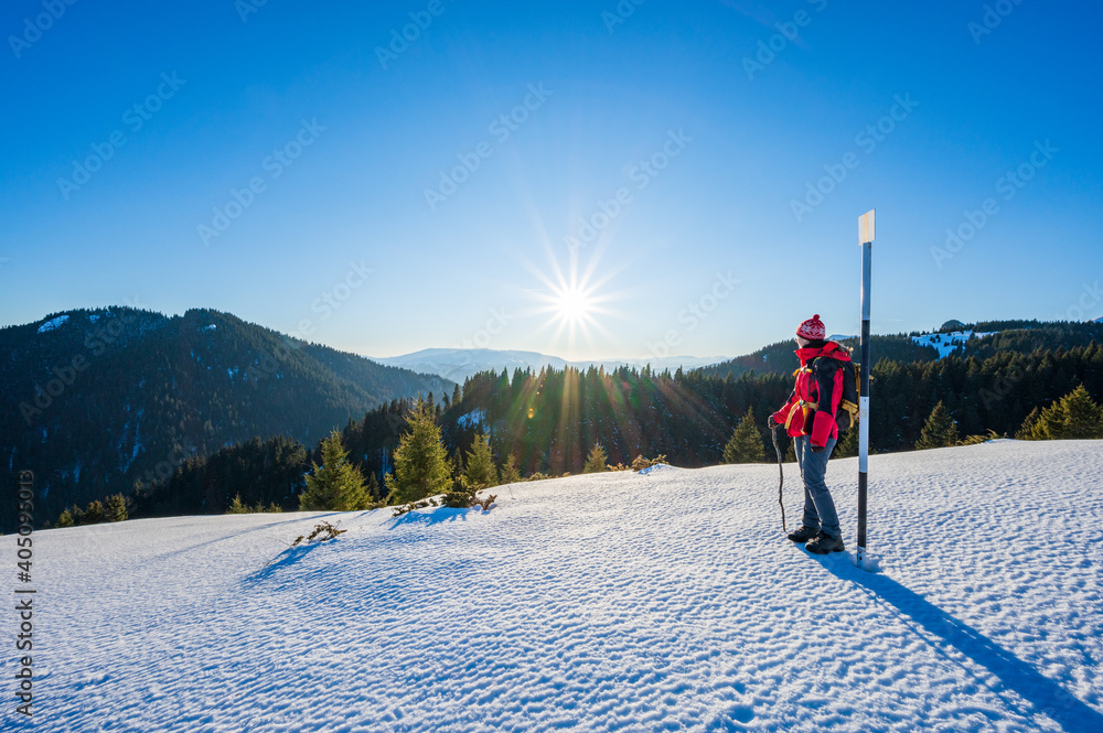 Female hiker standing next to trail sign looking over sunny mountain landscape in winter