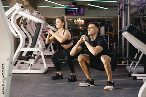 Two athletes perform squats. The coach shows the correctness of performing the exercises on the feet. Exercise in the gym. Joint training in small groups. A man and a girl train together.