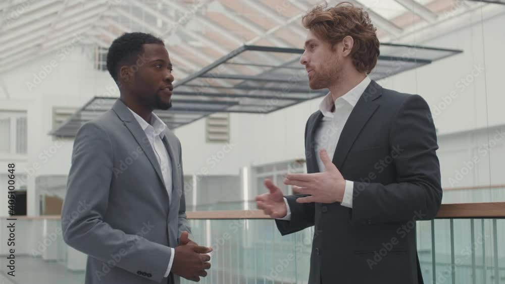 Lockdown of two diverse businessmen wearing suits standing in business center hall and chatting Lockdown of two diverse businessmen wearing suits standing in business center hall and chatting