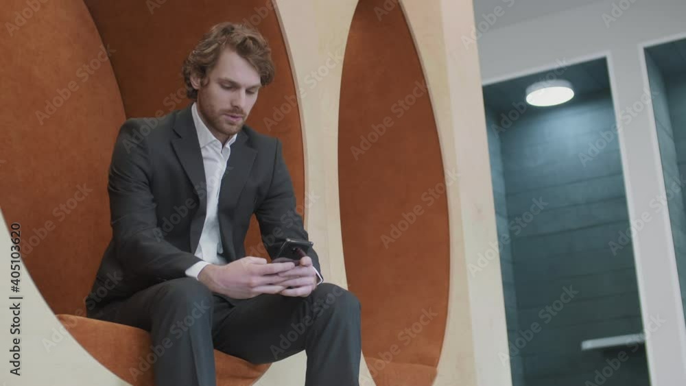 Medium shot of handsome ginger Caucasian businessman wearing suit sitting in hall in office and using smartphone Medium shot of handsome ginger Caucasian businessman wearing suit sitting in hall in office and using smartphone