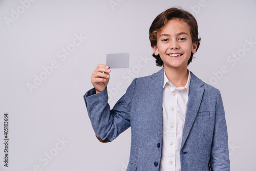 Cheerful small male kid holding credit card in formal outfit isolated over grey background