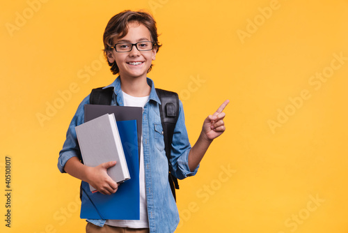 Smiling little boy pointing at copy space in casual clothes with books for studing at school isolated over yellow background photo