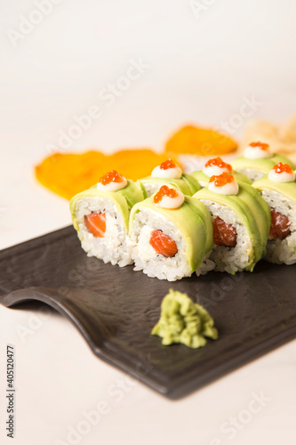 Close up to Japanese sushi roll with avocado and red caviar on top served with wasabi on black plate on white marble background. Raw Salmon, cream cheese wrapped in rice and nori seaweed. Inside out 