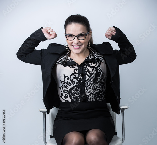 Donna d'affari bianca con i capelli raccolti e gli occhiali da vista, vestita con tailleur nero e camicia fantasia, esulta in segno di vittoria seduta nella sua poltrona, isolata su sfondo bianco photo