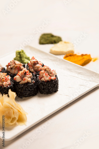 Side view of Japanese black rice sushi roll with unagi eel served with ginger  wasabi on square plate. Heart shapes if basil  garlic  turmeric powder spices on white marble background 