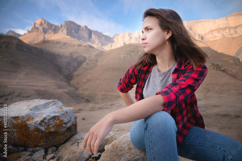 Naklejka premium Attractive brooding young caucasian woman in gray T-shirt and jeans sits on a cliff against the mountains on a sunny day