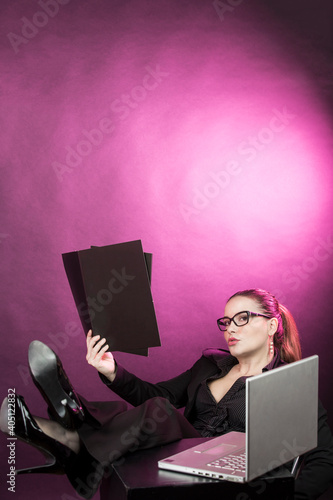 Manager donna con i capelli raccolti  con occhiali da vista, vestita con  tailleur nero lavora al computer con i piedi sopra il tavolo , isolata su sfondo rosso photo