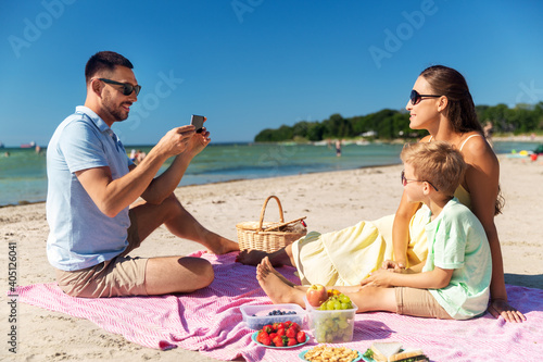 family, leisure and people concept - happy father with smartphone photographing mother and son on summer beach