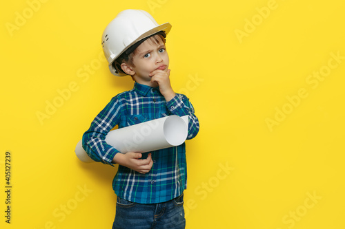 Serious little kid boy engineer or architect in a protective helmet holding construction plan over yellow background.