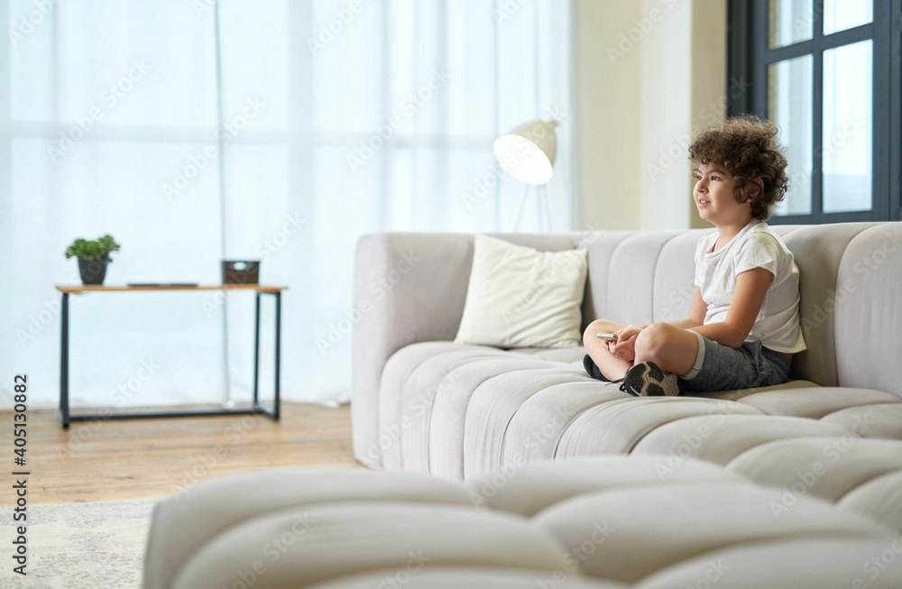 Adorable latin boy sitting on a sofa with smartphone and watching tv while spending time at home Adorable latin boy sitting on a sofa with smartphone and watching tv while spending time at home