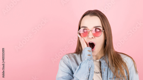 An excited girl in a denim shirt, holding her head in her hands, screamed and looked away. A three-quarter-length Studio shot on a pink background. © MP Studio