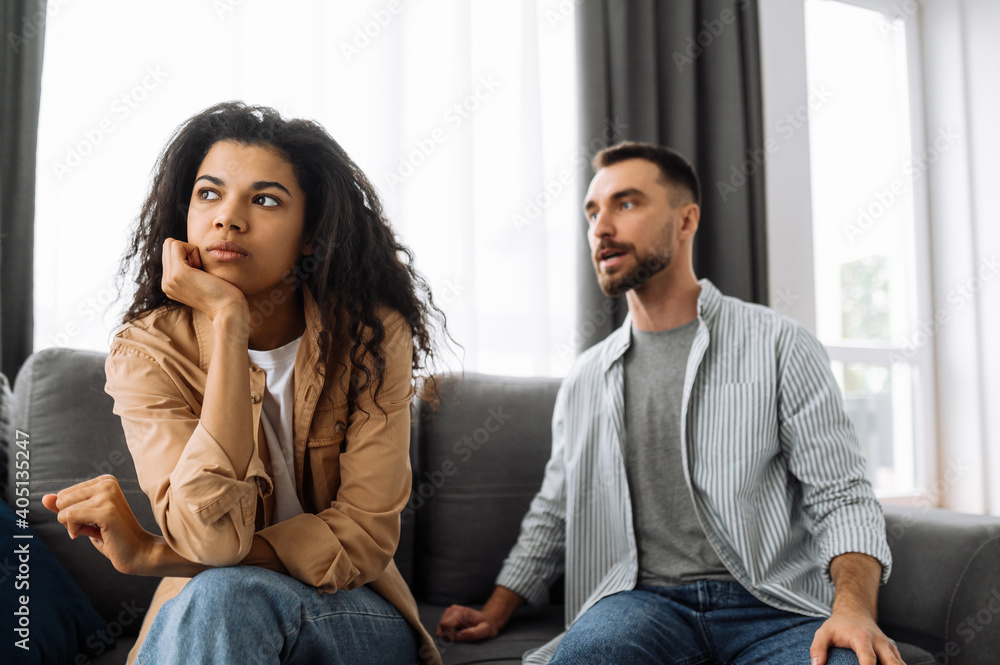 Multiracial couple in love quarreling at home on couch over household or financial problems. The woman is offended by the man and ignores him. Domestic family conflicts concept Multiracial couple in love quarreling at home on couch over household or financial problems. The woman is offended by the man and ignores him. Domestic family conflicts concept