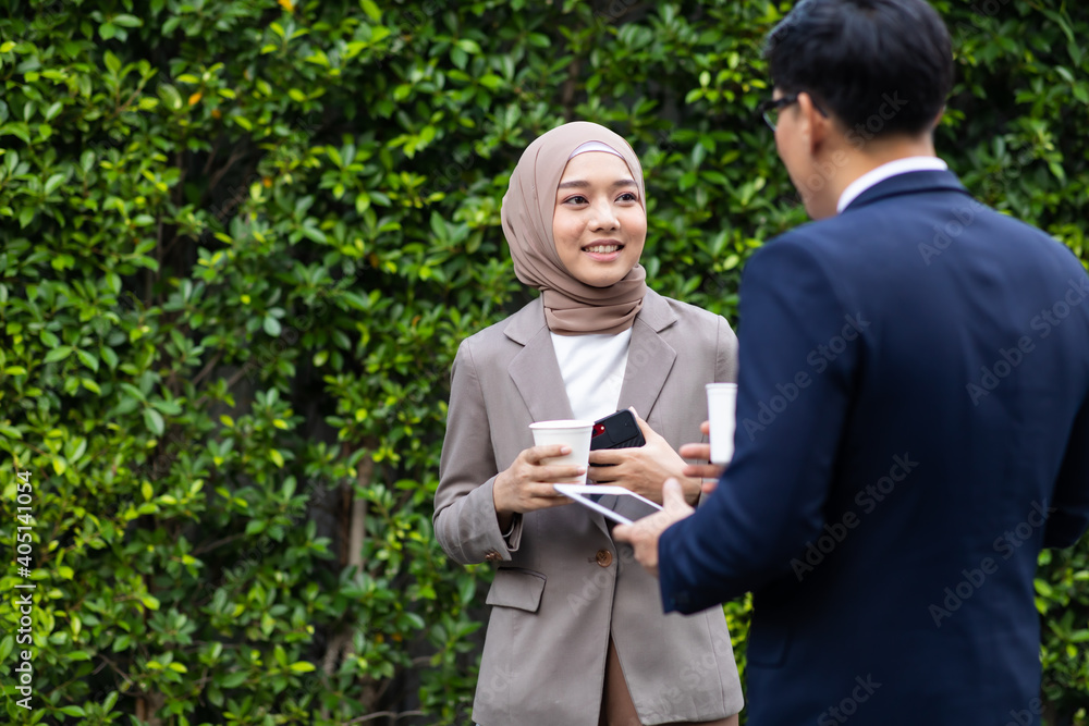 Young business muslim woman and business man colleague walking and discussing outside the office. Woman is holding a coffee and businessman is holding a digital tablet outdoor. Young business muslim woman and business man colleague walking and discussing outside the office. Woman is holding a coffee and businessman is holding a digital tablet outdoor.