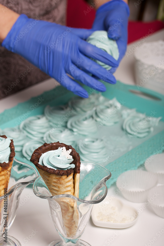 Woman in rubber gloves makes marshmallows. Nearby is a marshmallow in a waffle cone. Dessert lies on the table surface.