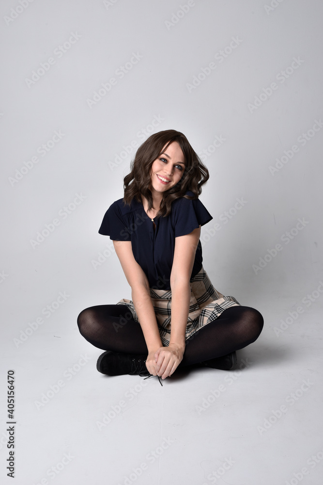 Full length portrait of pretty brunette woman wearing tartan skirt and blouse. Sitting pose on the ground, against a studio background. Full length portrait of pretty brunette woman wearing tartan skirt and blouse. Sitting pose on the ground, against a studio background.