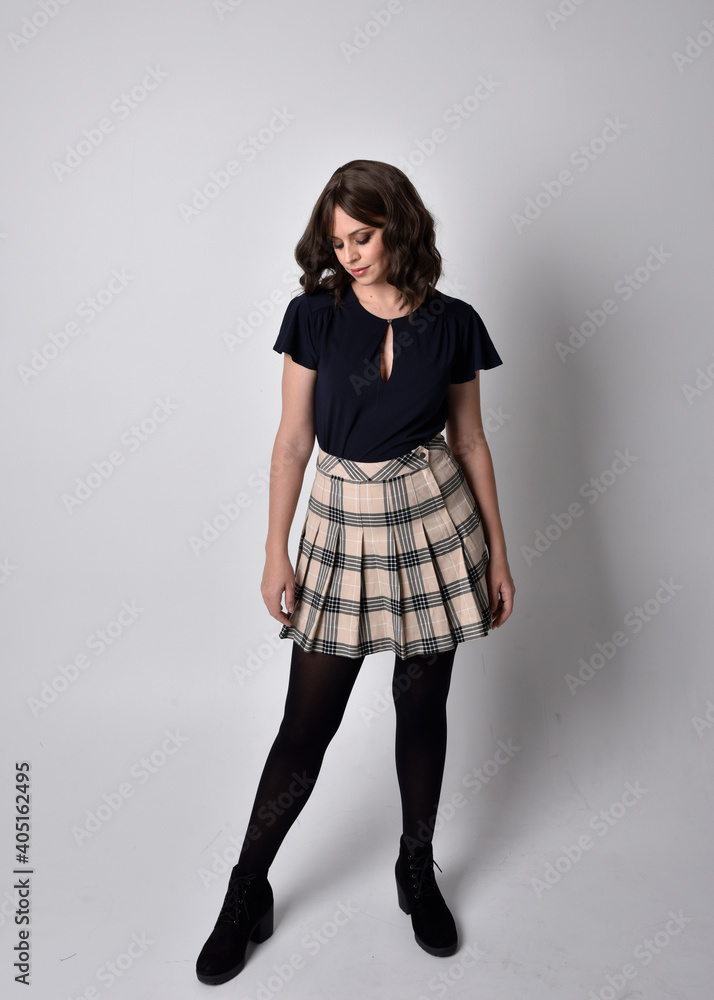 Full length portrait of pretty brunette woman wearing tartan skirt and blouse. Standing pose on the ground, against a studio background. Full length portrait of pretty brunette woman wearing tartan skirt and blouse. Standing pose on the ground, against a studio background.