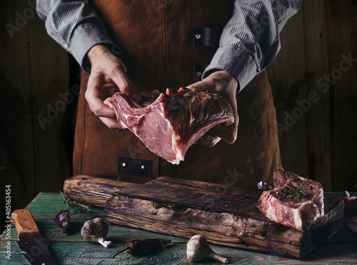 A man in a leather apron holds a piece of raw meat with a bone. Pork ribs in the hands of the butcher..