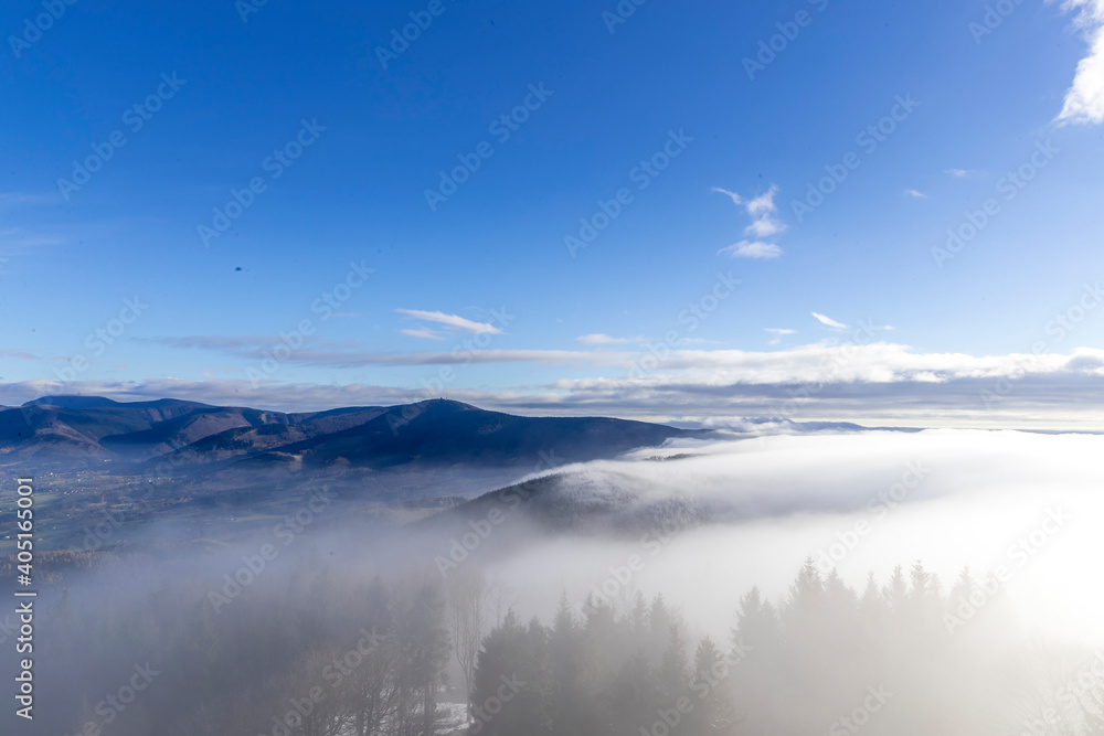 The tops of the hills during a sunny afternoon covered with thick fog which passes between the treetops in the background of blue sky.