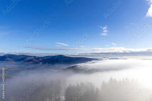 The tops of the hills during a sunny afternoon covered with thick fog which passes between the treetops in the background of blue sky. © Lukas