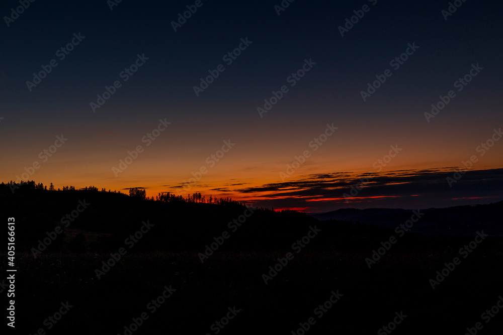 Field where grass grows overlooking the surrounding hills and treetops during sunset on an orange horizon overlooking the landscape and nature.