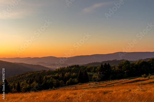 Sun setting over a horizon of orange hills with a coniferous tree on the left and clouds moving in the background over the Beskydy countryside. © Lukas