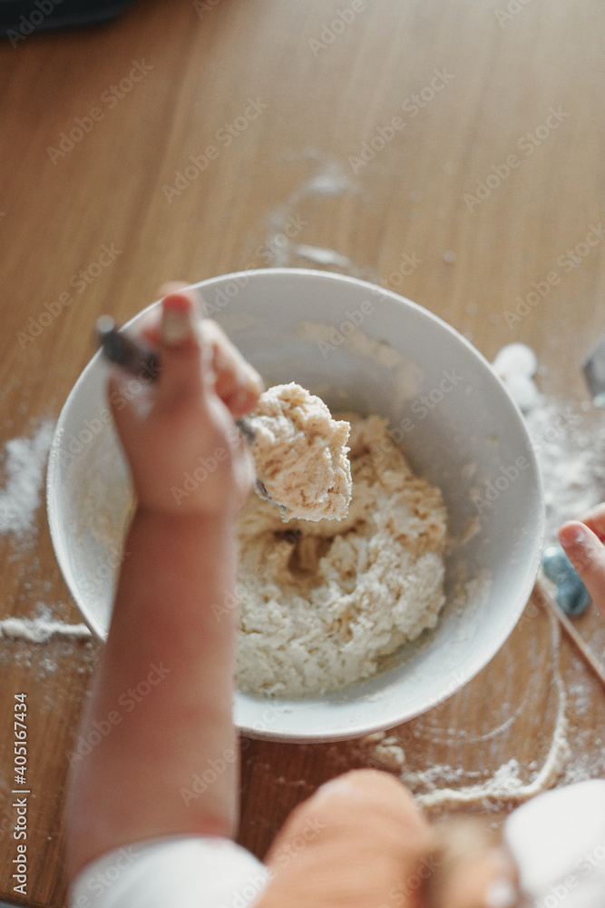hands of children stir a dough in the white bowl with spoon, on the wooden table in the kitchen. The process of mixing ingredients for pastry. Closeup, selective focus. hands of children stir a dough in the white bowl with spoon, on the wooden table in the kitchen. The process of mixing ingredients for pastry. Closeup, selective focus.