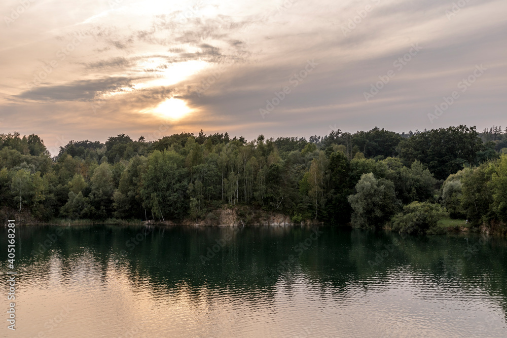 Flooded quarry and trees lying on the shore during sunset with fast moving clouds and a blue-orange sky in the background.