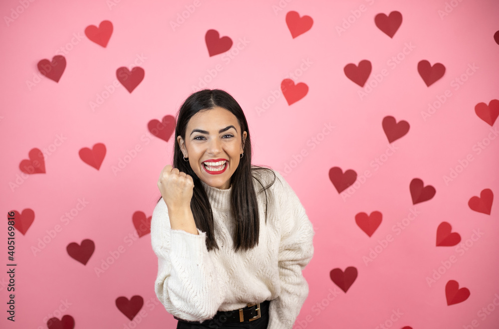 Young beautiful woman over pink background with harts angry and mad raising fist frustrated and furious while shouting with anger. Rage and aggressive concept. Young beautiful woman over pink background with harts angry and mad raising fist frustrated and furious while shouting with anger. Rage and aggressive concept.