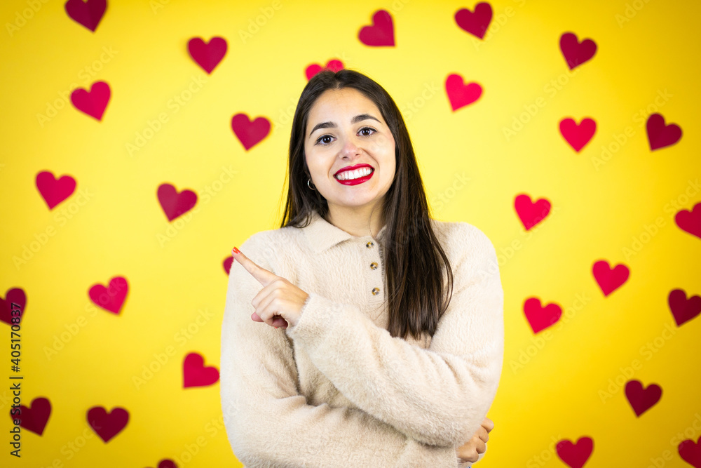 Young caucasian woman over yellow background with red hearts smiling and pointing with hand and finger to the side Young caucasian woman over yellow background with red hearts smiling and pointing with hand and finger to the side