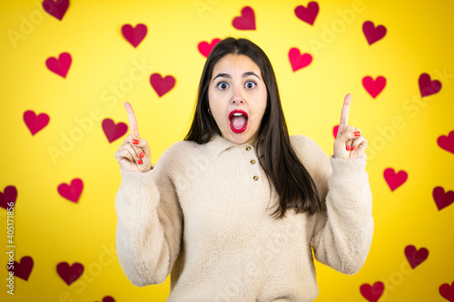 Young caucasian woman over yellow background with red hearts amazed and surprised looking at the camera and pointing up with fingers and raised arms