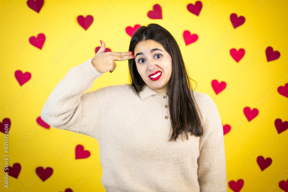 Young caucasian woman over yellow background with red hearts Shooting and killing oneself pointing hand and fingers to head like gun, suicide gesture. Young caucasian woman over yellow background with red hearts Shooting and killing oneself pointing hand and fingers to head like gun, suicide gesture.