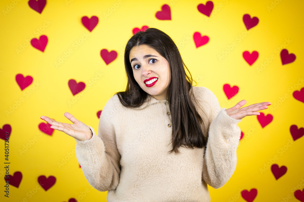 Young caucasian woman over yellow background with red hearts clueless and confused expression with arms and hands raised Young caucasian woman over yellow background with red hearts clueless and confused expression with arms and hands raised