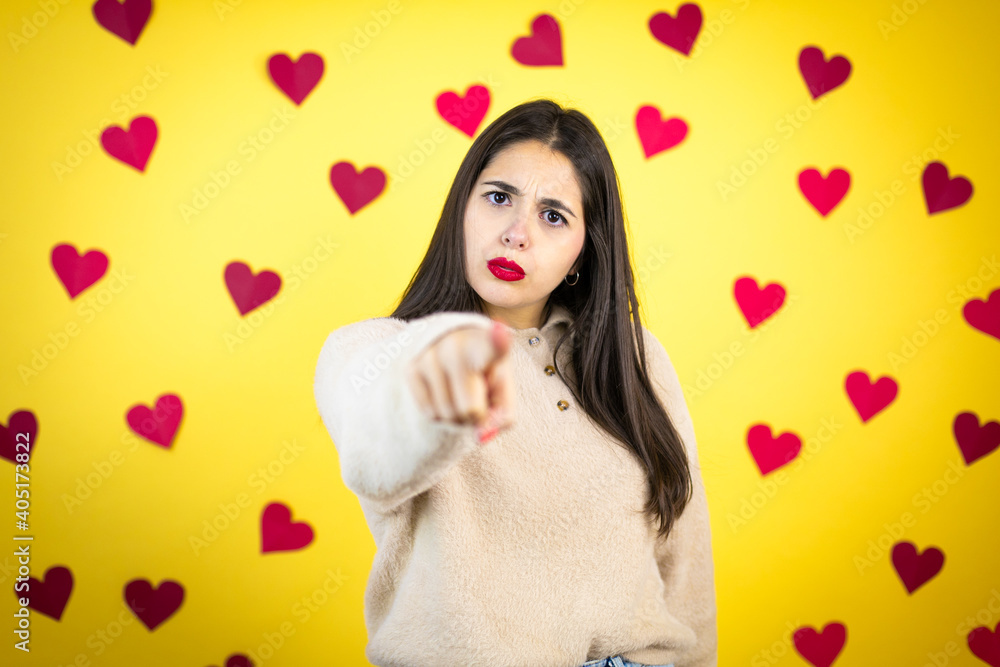 Young caucasian woman over yellow background with red hearts pointing with finger to the camera and to you, confident gesture looking serious Young caucasian woman over yellow background with red hearts pointing with finger to the camera and to you, confident gesture looking serious