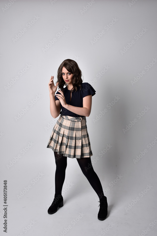 Full length portrait of pretty brunette woman wearing tartan skirt and blouse.  Standing pose on the ground,  against a  studio background.