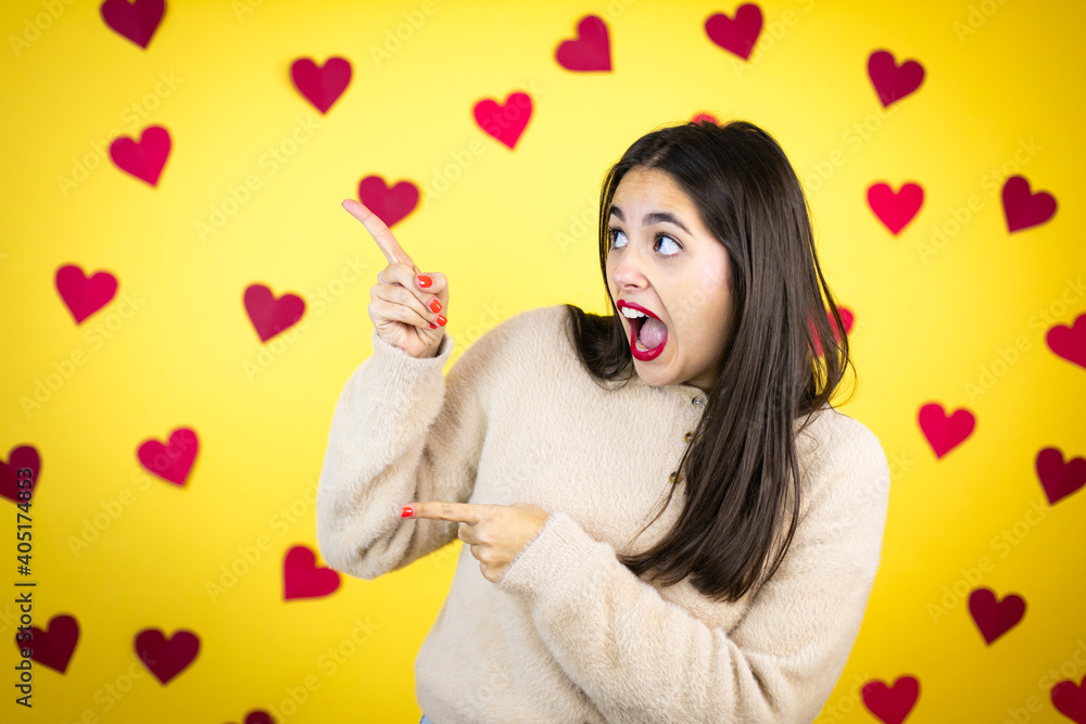 Young caucasian woman over yellow background with red hearts surprised and pointing her fingers side Young caucasian woman over yellow background with red hearts surprised and pointing her fingers side