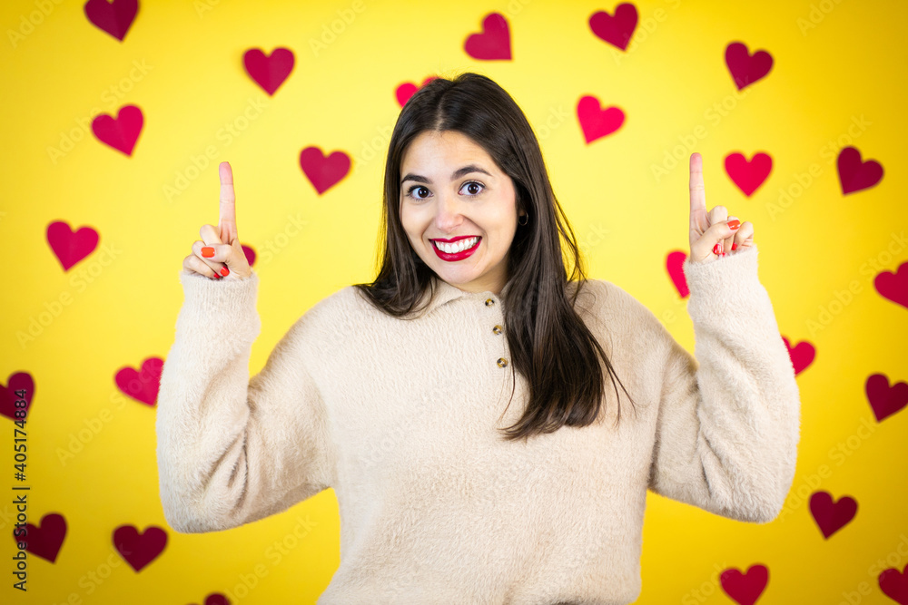 Young caucasian woman over yellow background with red hearts smiling, looking at the camera and pointing up with fingers and raised arms Young caucasian woman over yellow background with red hearts smiling, looking at the camera and pointing up with fingers and raised arms