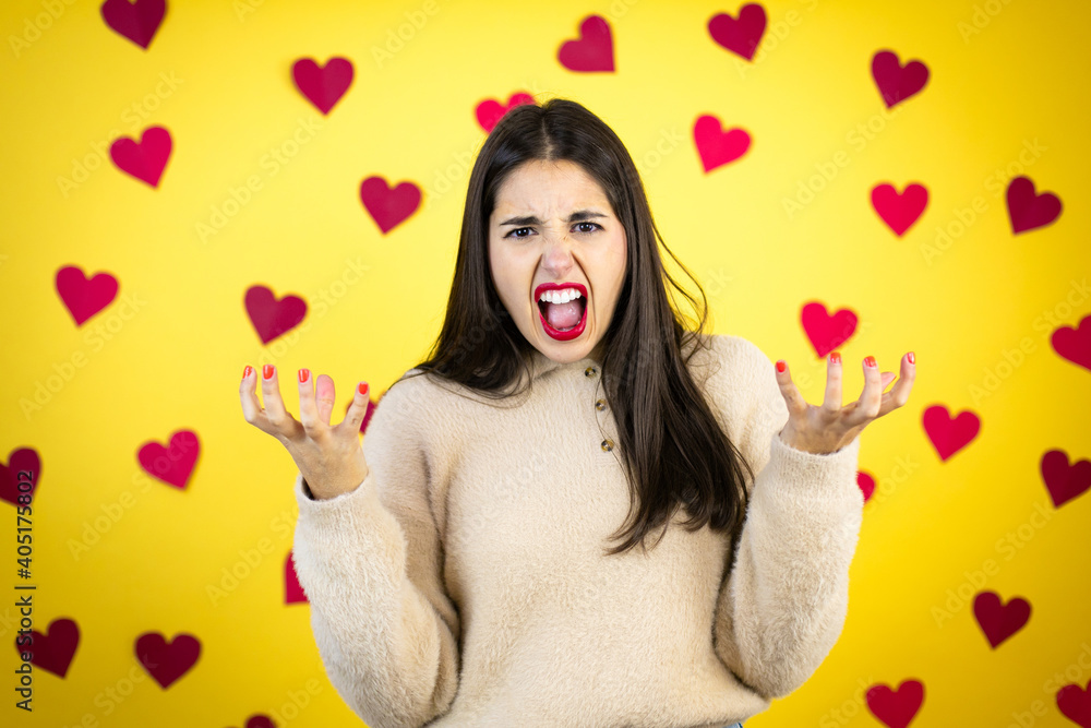 Young caucasian woman over yellow background with red hearts crazy and mad shouting and yelling with aggressive expression and arms raised. Frustration concept. Young caucasian woman over yellow background with red hearts crazy and mad shouting and yelling with aggressive expression and arms raised. Frustration concept.