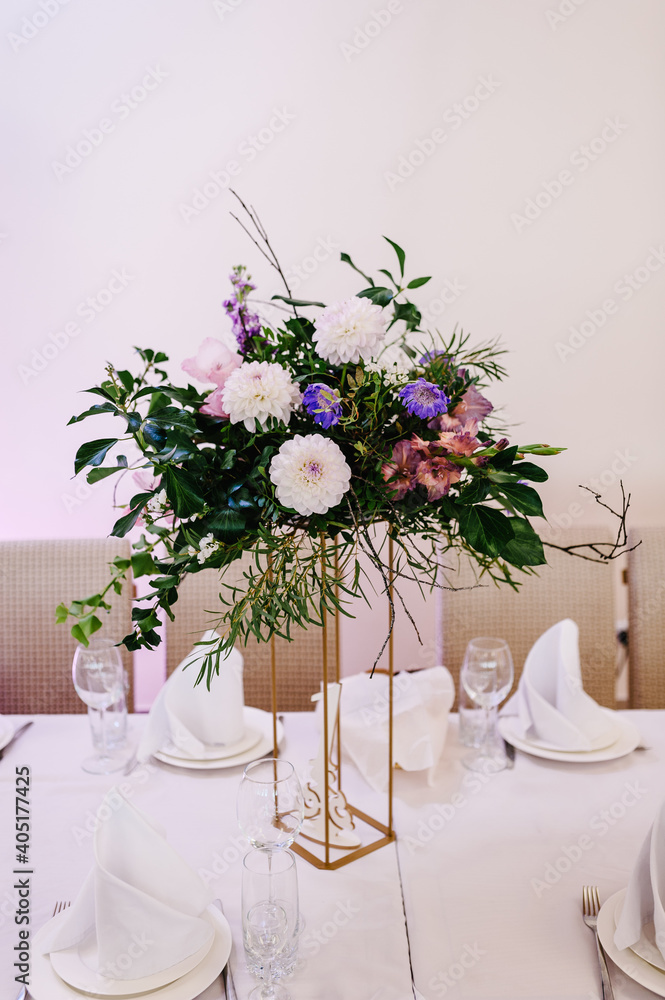 Table covered with a tablecloth and served with dishes and cutlery. Festive table decorated with composition of flowers and greenery in the wedding banquet hall.