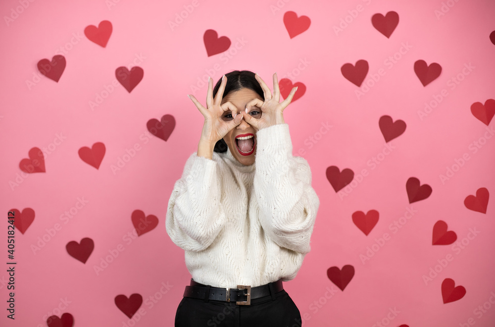 Young beautiful woman over pink background with harts doing ok gesture shocked with smiling face, eye looking through fingers Young beautiful woman over pink background with harts doing ok gesture shocked with smiling face, eye looking through fingers