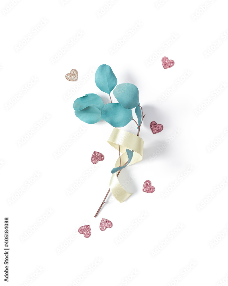 Fototapeta premium The eucalyptus branch is wrapped in a yellow satin ribbon and is on a white background. Shiny figures of hearts are scattered nearby. Valentine's Day minimalist layout. Flat lay. Top view.