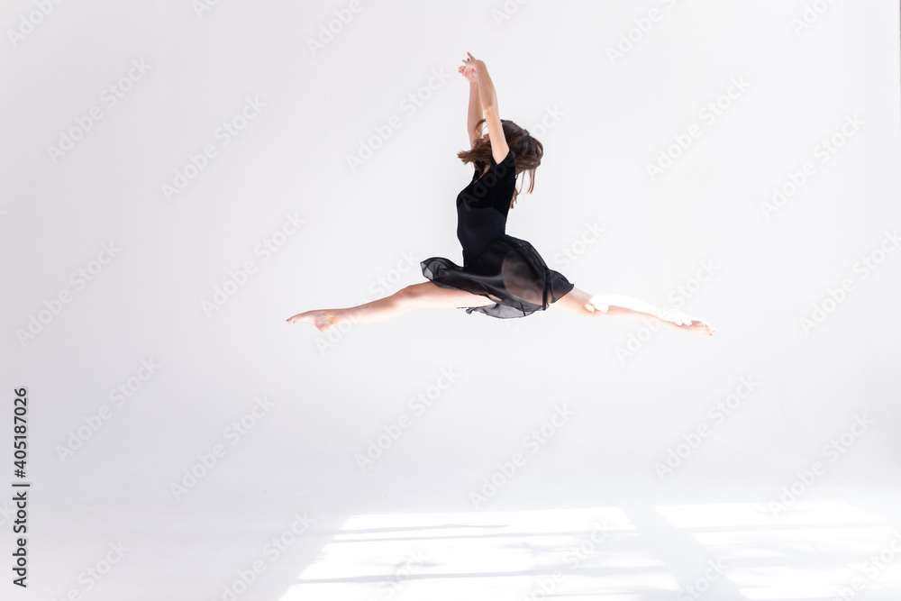A slender girl in a black sports swimsuit does a split in a jump on a white isolated background. Girl athlete doing splits in the air on a white isolated background with place for text or logo.