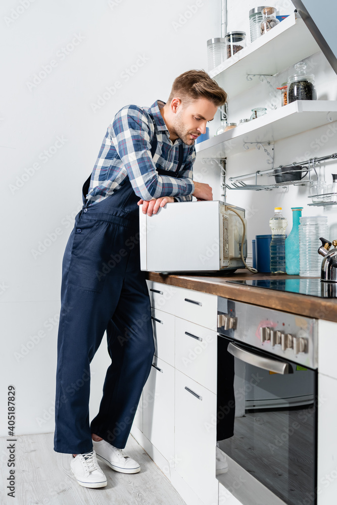 full length of young repairman looking at microwave oven in kitchen