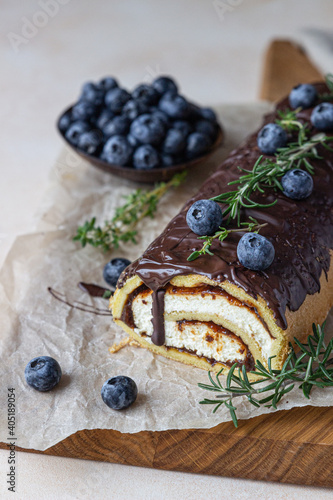 Sponge cake roll with chocolate and cream cheese decorated with chocolate glaze, blueberry and rosemary on parchment, light background. Biscuit swiss roll.